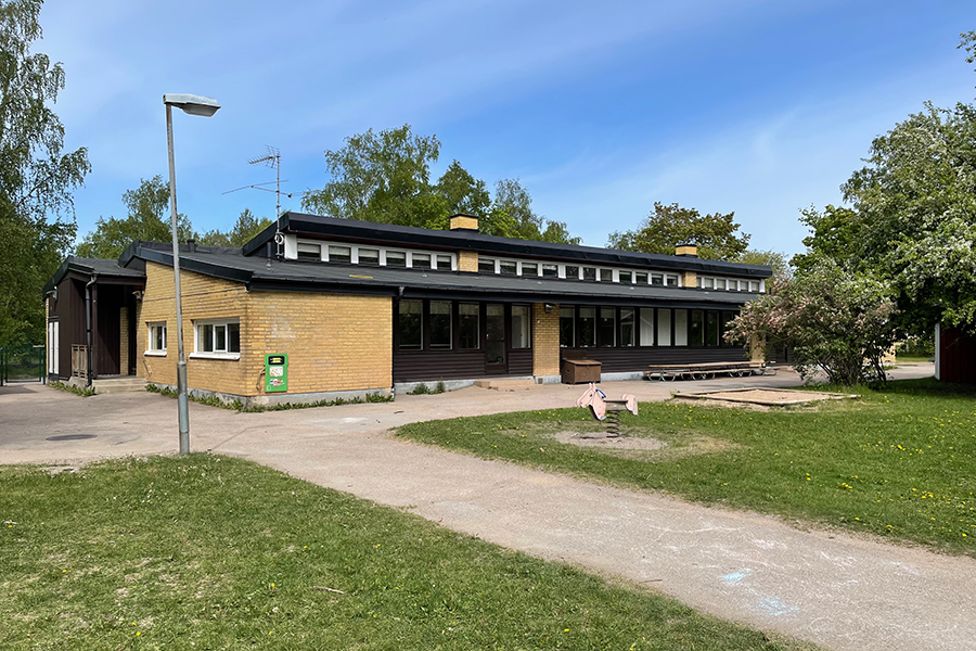 Mårbackas förskola. n gul tegelbyggnad med svart tak och svarta trädetaljer. Grönyta runt om byggnaden. 