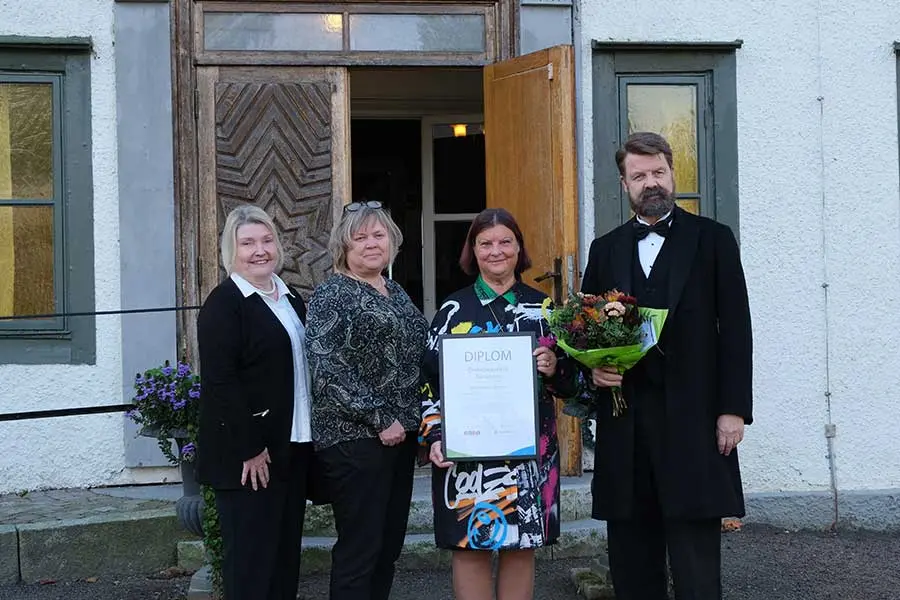 Fyra personer står framför en byggning, varav en håller ett blomsterbukett och en visar ett diplom med texten "DIPLOM".