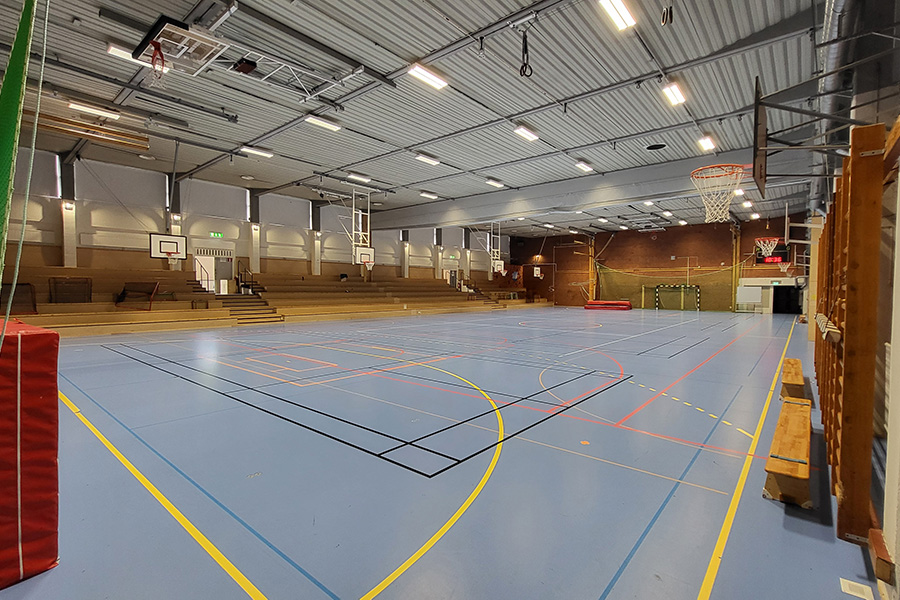 Idrottssal med blå golvbeläggning. Det finns basketkorgar, handbollsmål och tjockmattor. På sidorna finns sittplatser, bänkar och ribbstolar längs väggarna. 