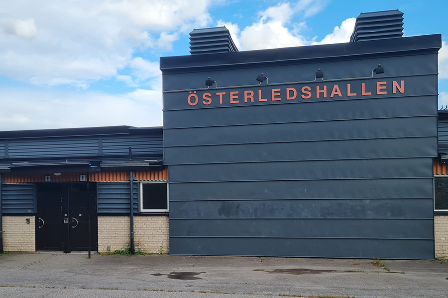 Entrédörrar till skolbyggnad med fasad i vitt tegel och blått trä. Stor skylt med text Öserledshallen. Asfalterad yta framför.