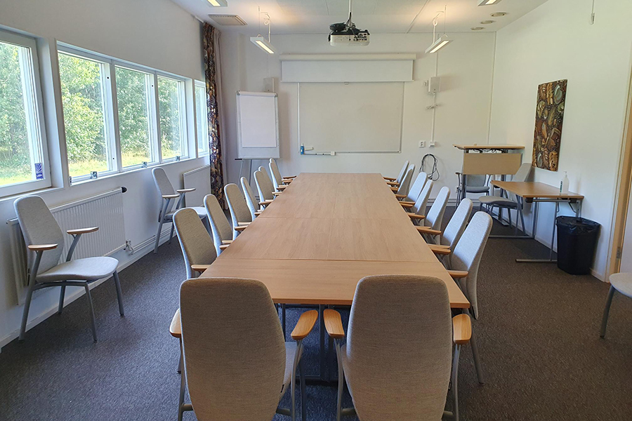 Möteslokal med bord och stolar. Whiteboardtavla på stativ och på vägg. Projektor i taket. Längs ena väggen finns talarpulpet och extrabord och stolar.