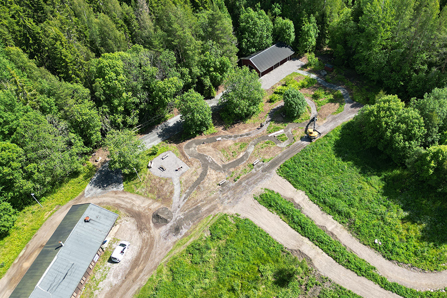 En bild tagen från luften. En maskin står på en grusad yta, runtomkring finns skog och mindre byggnader.