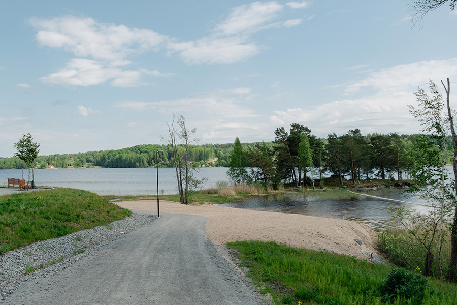 En sandstrand intill vattnet. I mitten av bilden finns en udde med många träd på, som går ut i vattnet