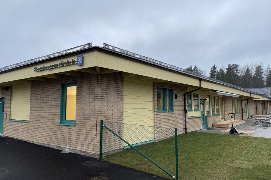 Ett hus med gult tegel och gröna brädor runt fönstren. Till höger skymtas en utegård med lekutrustning. 