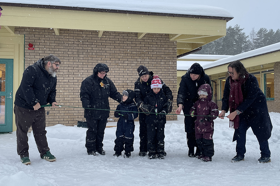 Tre barn och fem vuxna står på rad. Barnen håller i en varsin sax och hjälps åt att klippa i ett grönt band som hålls utsträckt framför dem. 