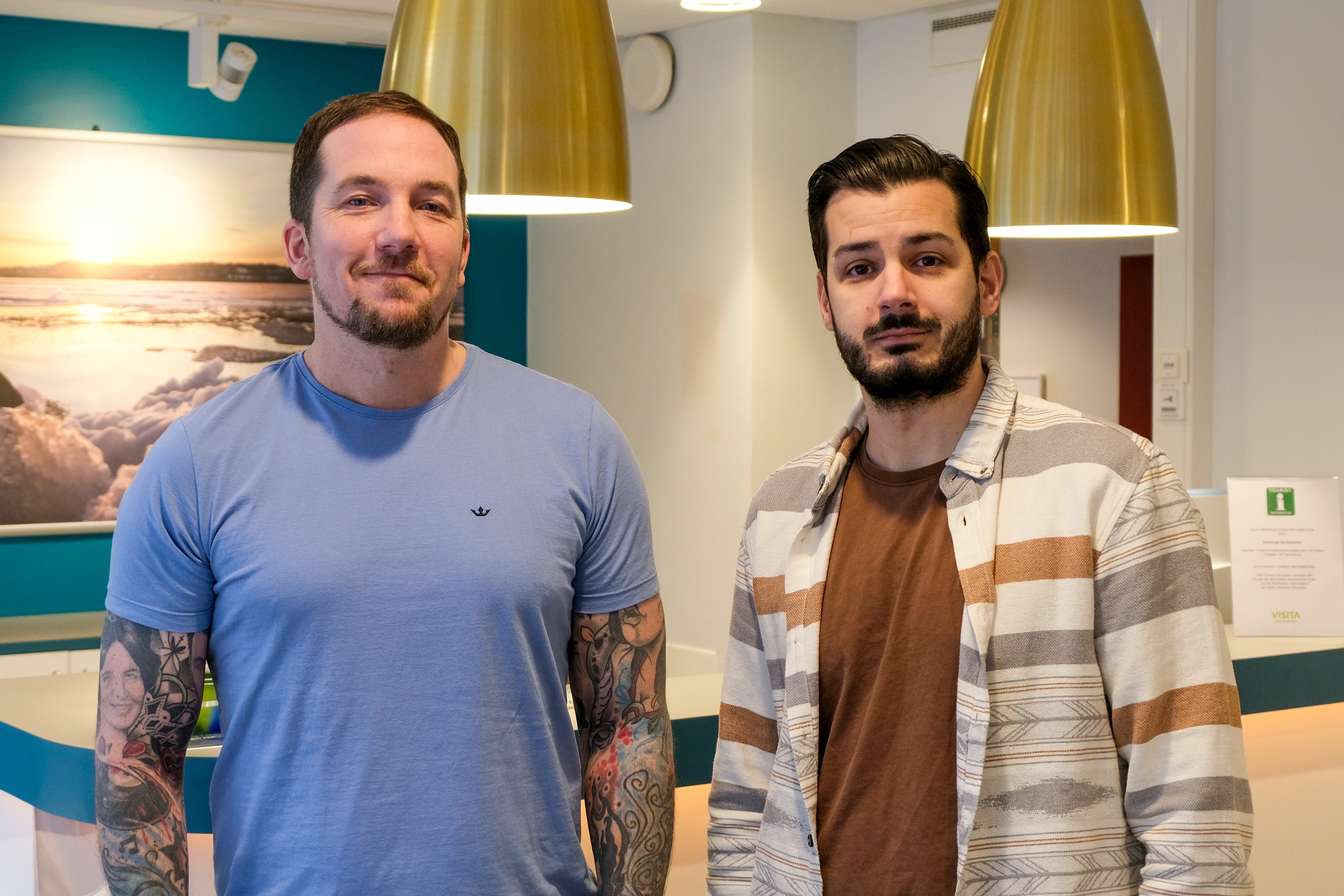 Två män står vid en servicedisk med stora guldfärgade lampor som hänger från taket. Den ena mannen har kort mörkt hår, tatueringar och en blå T-shirt. Den andra mannen har mörkt hår och skägg och bär en brun T-shirt och en vitmönstrad skjorta.