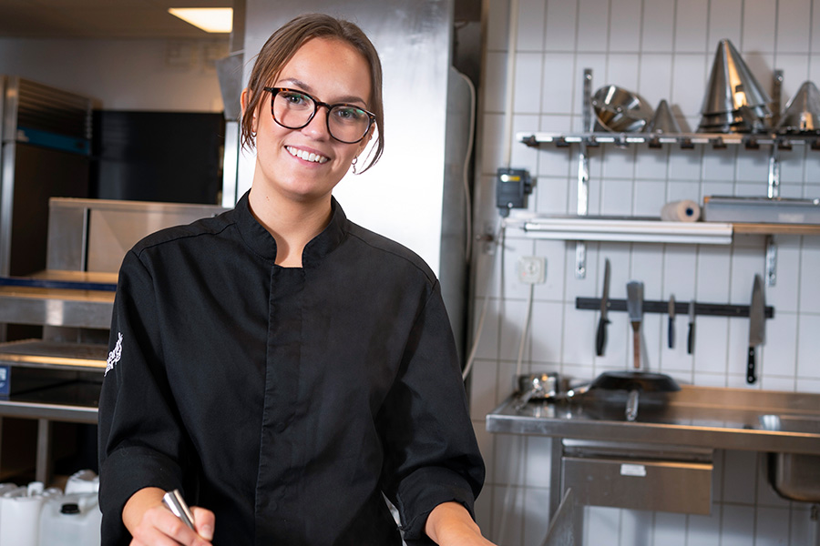 Elev står vid spisen i skolköket och ler in i kameran. Hon är klädd i kockkläder. 