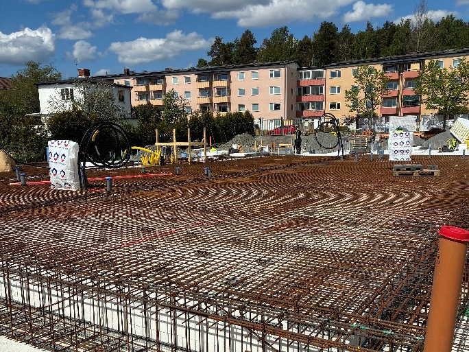 En byggarbetsplats med armeringsnät och en byggnad med flera våningar i bakgrunden. Himlen är blå med vita moln.