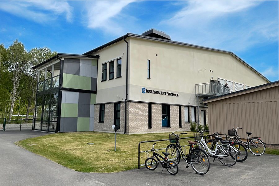 Bullerdalens förskola.Två våningshus i naturfärger med en glasentré och en grön ytan utanför. Cyklar står parkerade intill grönytan. 