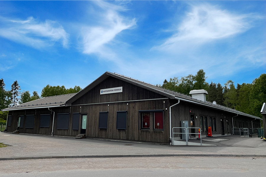 Kartografens förskola. En stor träbyggnad med spetsigt tak med asfalt runt byggnaden och blå himmel.