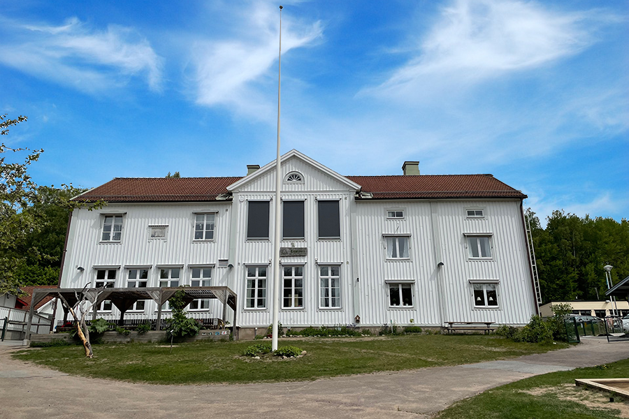 Sandmos förskola. En stor vit byggnad med ljusgråa knutar med grönyta framför byggnaden och en flaggstång.  