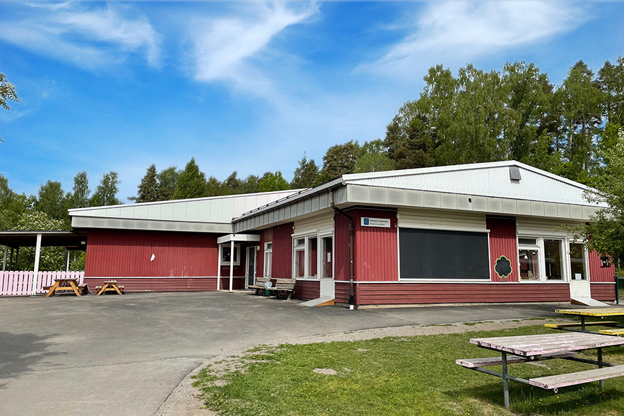 Smedens förskola. Röd byggnad med plåttak med asfalt utanför byggnaden och skogen i bakgrunden. 