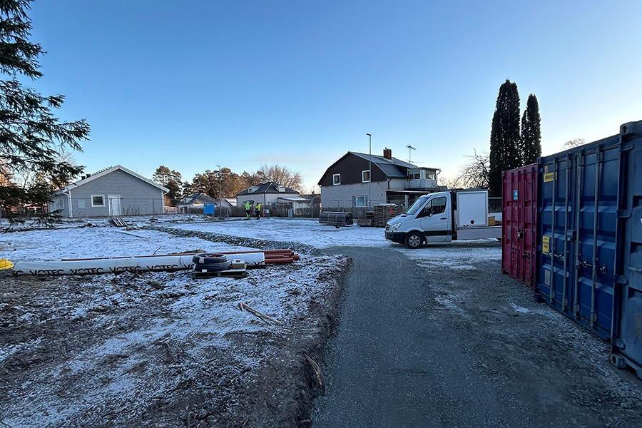 En öppen plan med några rör som ligger på marken. I bakgrunden syns några hus och till höger i bild står en röd och en blå container.