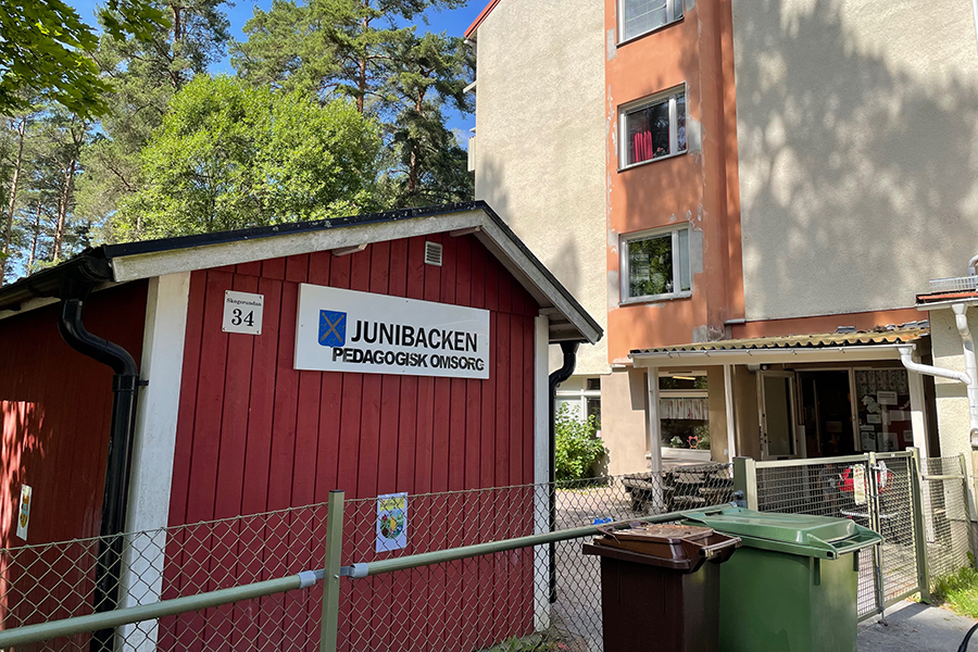 En liten röd stuga med grönt staket utanför och ett lägenhetshus bredvid.