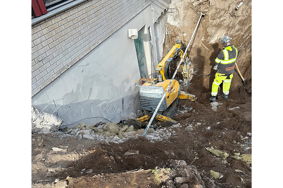En grop med grus intill en husfasad. I gropen står en gul maskin och en person iklädd arbetskläder.