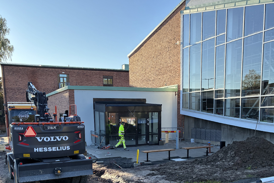 En byggnad med entrédörrar i glas. Till vänster står en grävmaskin.