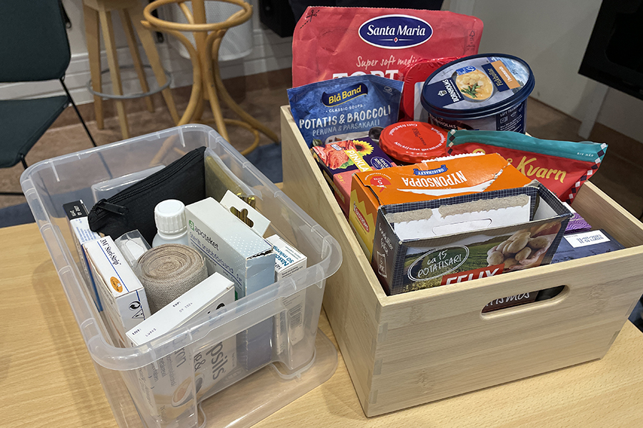 En plastlåda som står till vänster i bild innehållande olika förpackningar med sjukvårdsprodukter. Till höger står en trälåda innehållande olika förpackningar med matprodukter. 