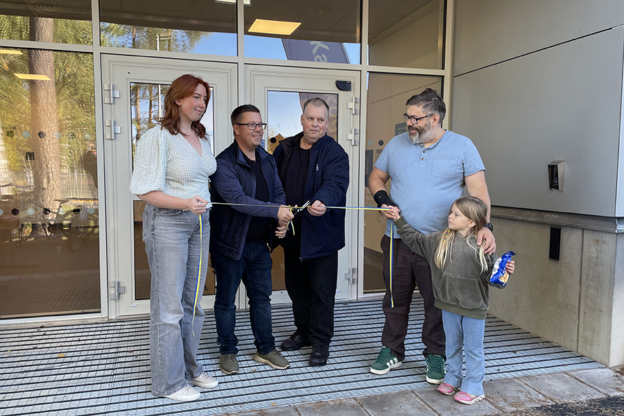 Fyra personer står vid en entré till en byggnad. Två av personerna håller i ett band som de andra två klipper itu med en sax. 