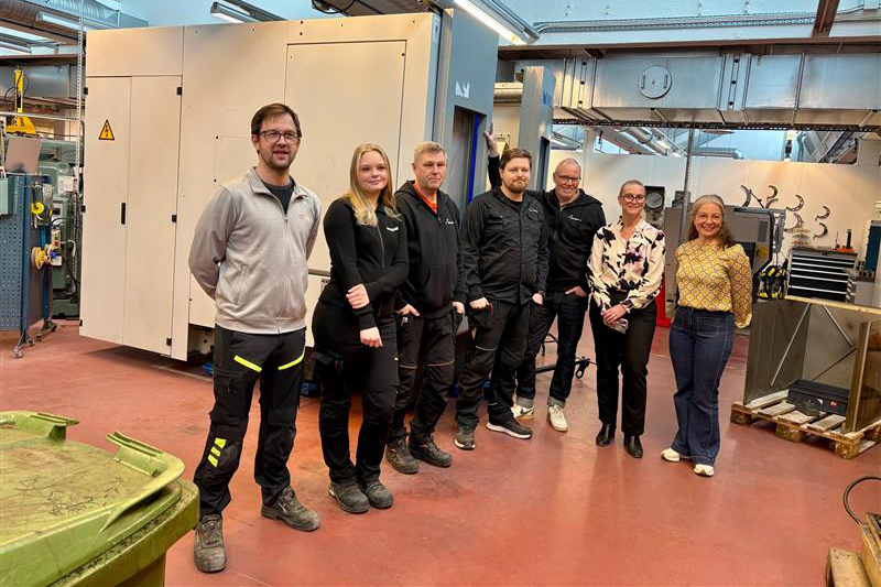 Sju personer, tre kvinnor och fyra män, står framför en maskin i storleken av en husvagn. Fyra av dem bär varselkläder och tre av dem är civilklädda.