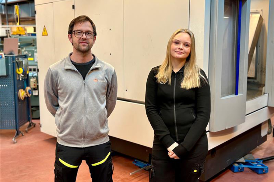 Två personer i varselkläder står framför en stor industrimaskin i form av en låda. Till vänster står en mörkhårig man med glasögon och grå hoodie och till höger står en ung, blond kvinna med svart hoodie.