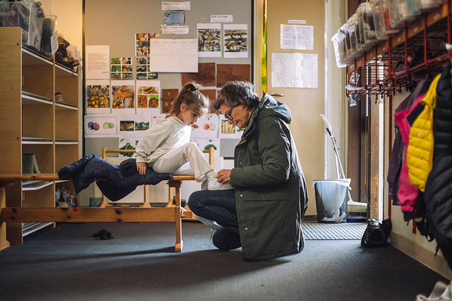En äldre kvinna hjälper en liten flicka att ta på en sko i kapprummet på en skola.