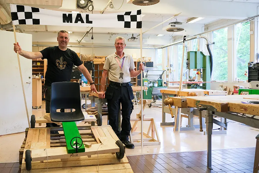 Två män står bredvid en lådbil och håller en banderoll med texten "MÅL" synlig ovanför. Verkstadsmiljö med flera arbetsbänkar i bakgrunden.