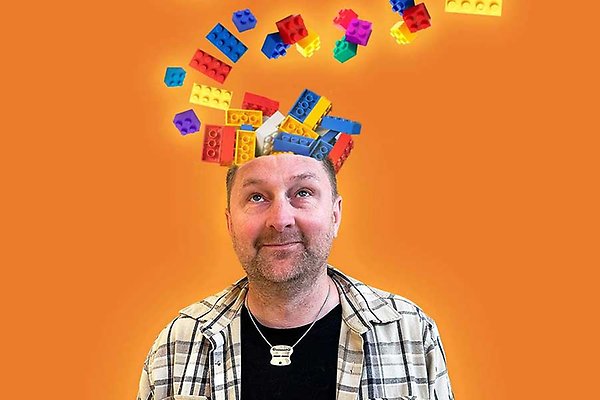 En man ler mot kameran med legobitar på huvudet. Legobitar i rött, blått och gult mot en orangefärgad bakgrund.