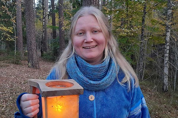 En person med ljusblont hår i en blåjacka håller en lykta i en skogsmiljö.