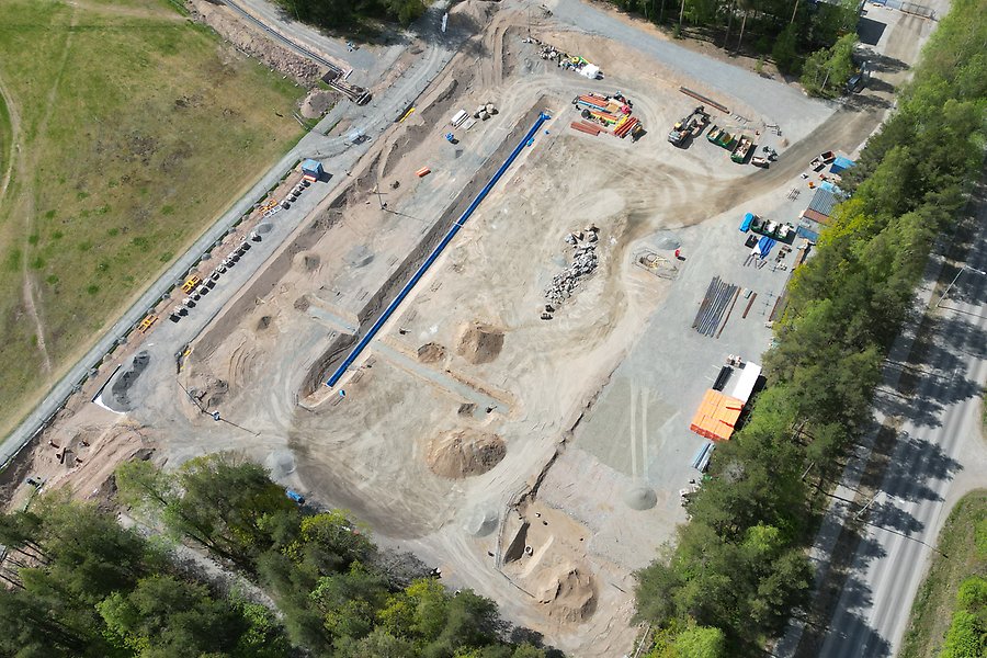 En bild tagen från luften över en byggarbetsplats. Ytan är täckt av grus och på sidan står en rad med containrar i olika färger.