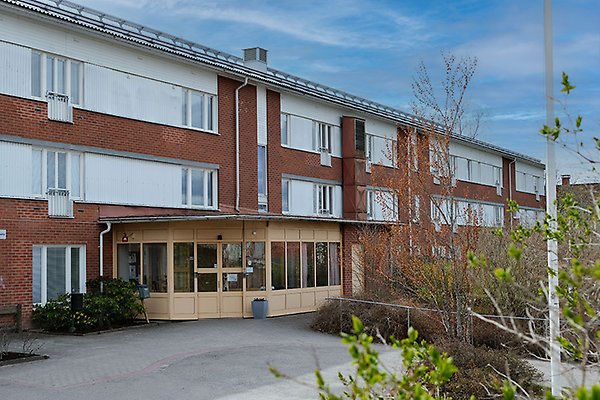 En tegelbyggnad med huskroppar och en entré. I förgrunden syns grönska. 